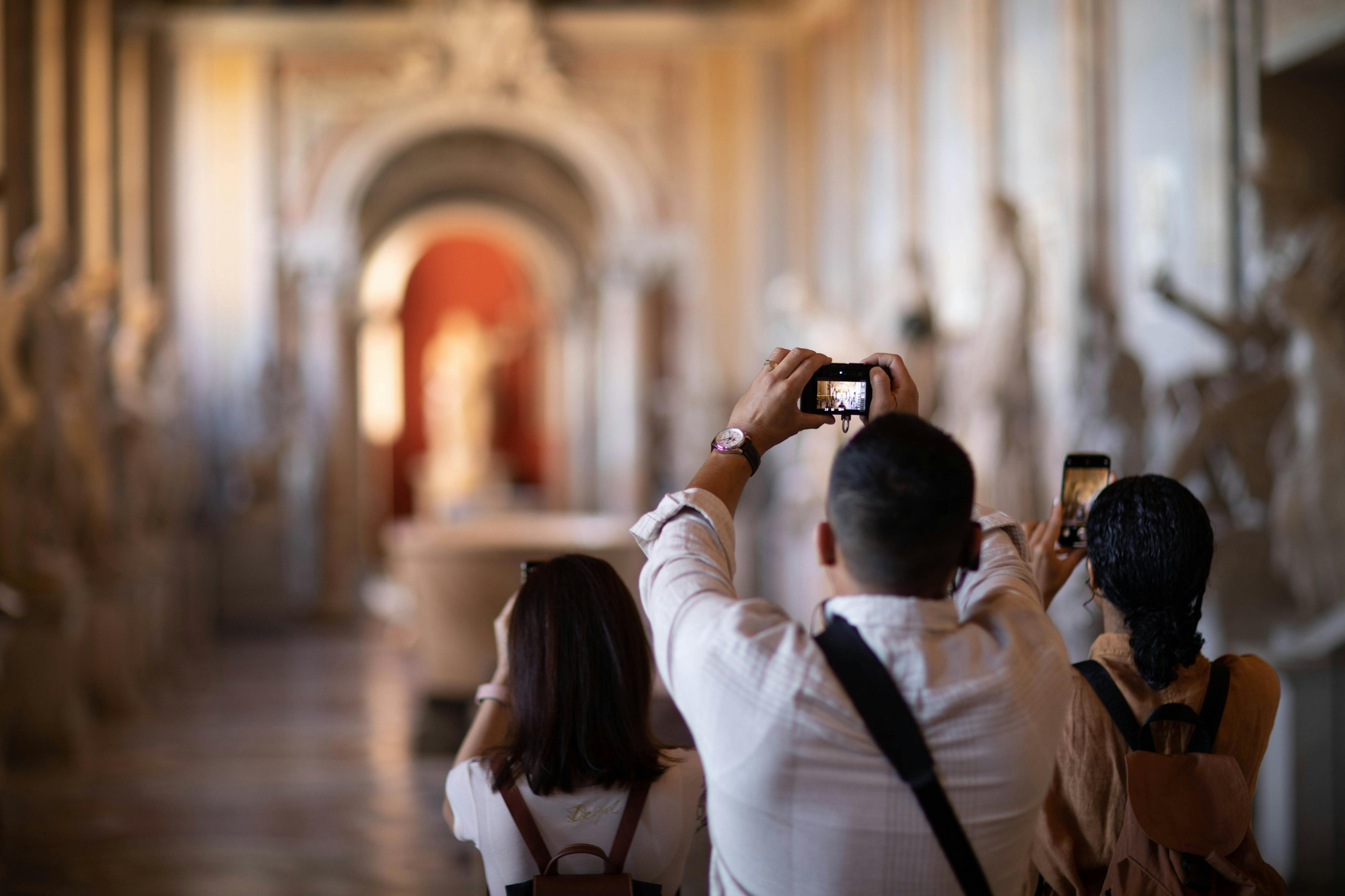 Persone che scattano foto in una sala di un museo, con delle sculture e una porta ad arco visibili sullo sfondo.