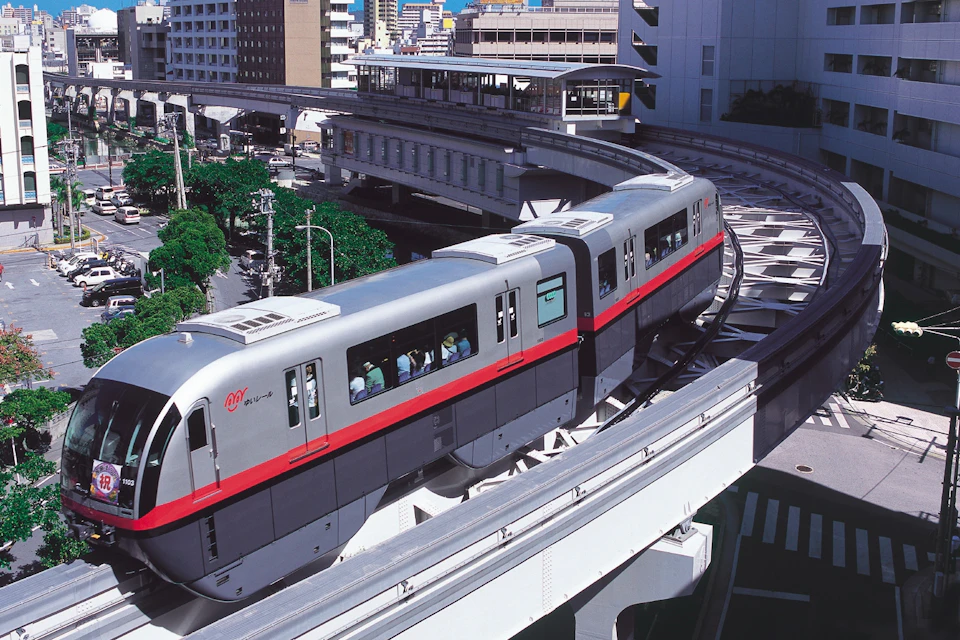 A modern monorail train traveling on elevated tracks in an urban area with surrounding buildings and green trees.