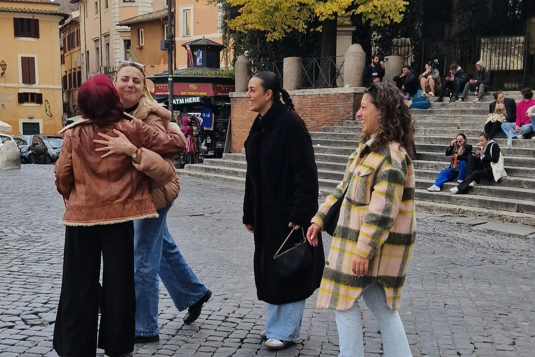 Two women hug warmly in a cobblestone square while another smiles nearby. Buildings and steps are visible in the background.