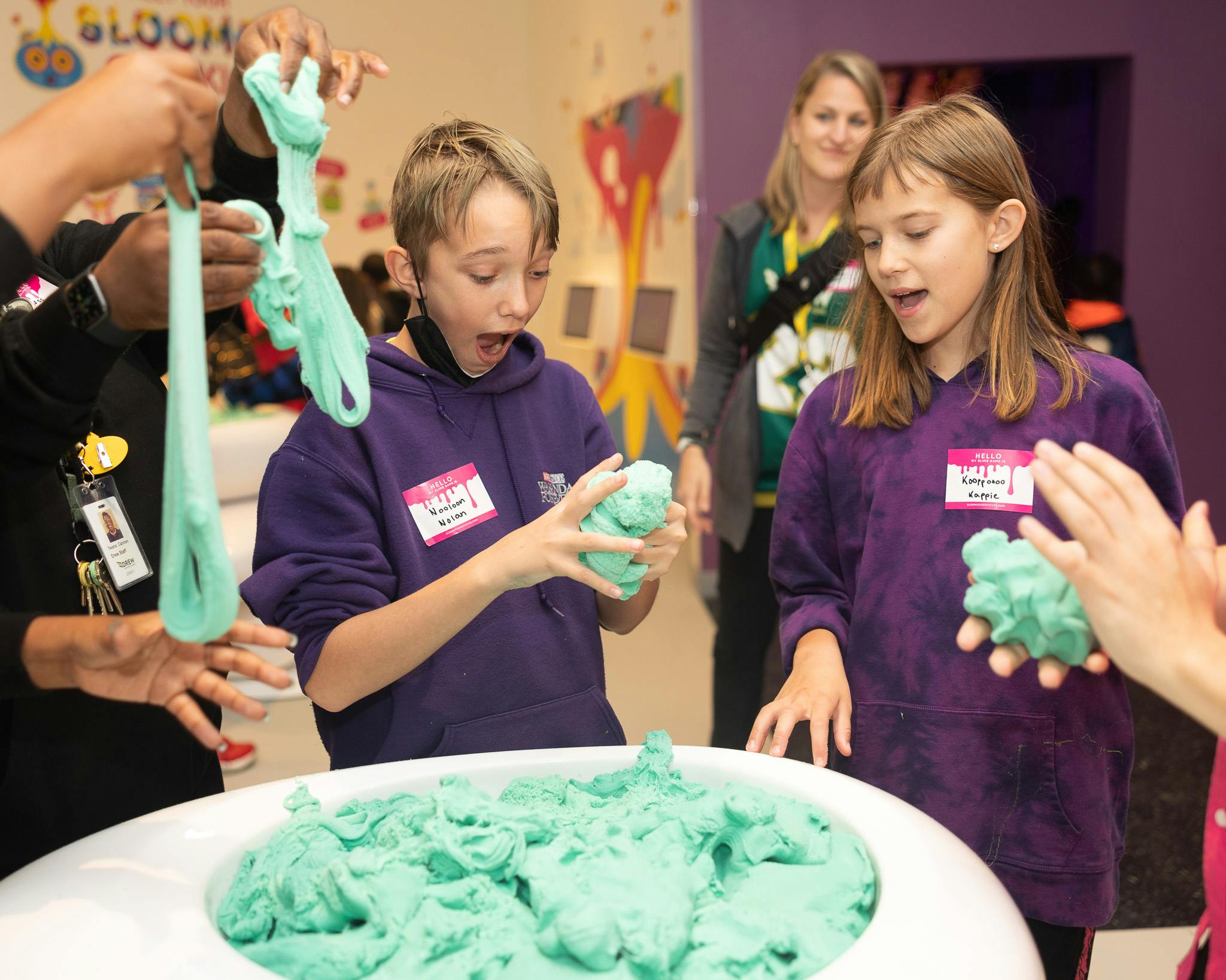 To børn i lilla hættetrøjer leger med grønt formbart skum ved et bord, under opsyn af en voksen i baggrunden.