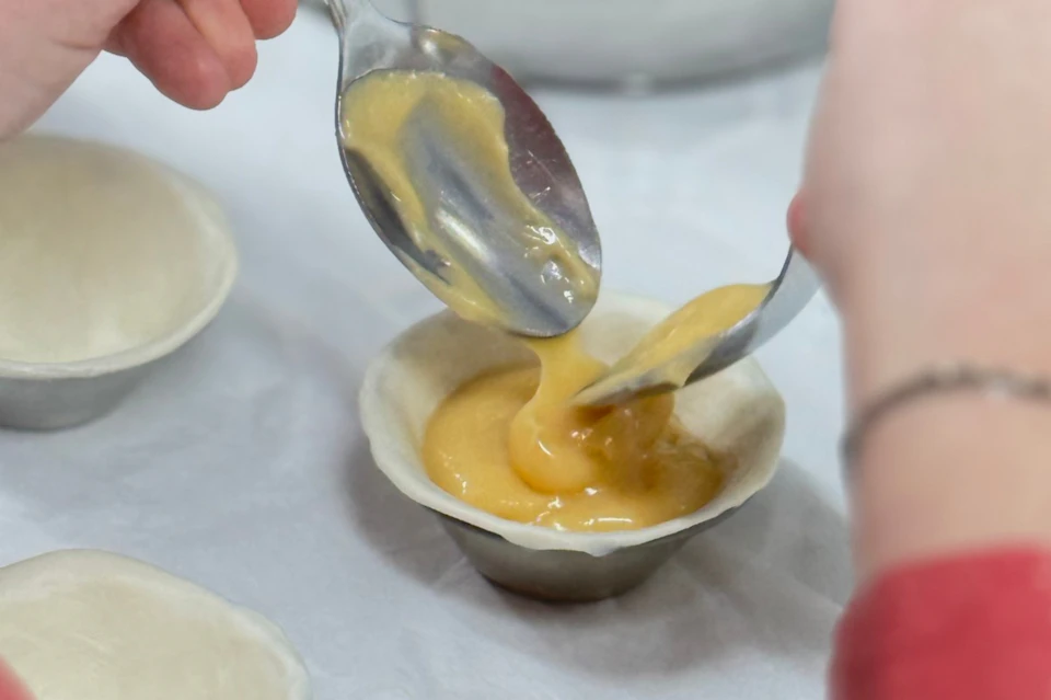 A person is filling a pastry shell with a yellow filling using spoons.