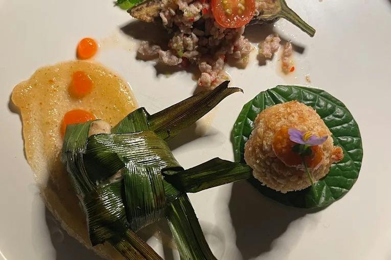 A plated dish featuring artistic food presentation with wrapped leaves, a round crispy item, and a vegetable garnish.