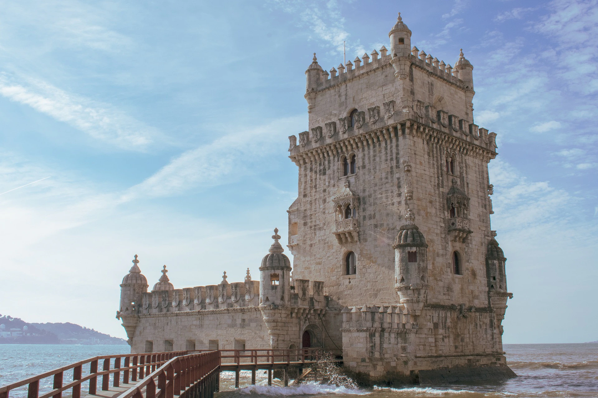 Historic stone tower with ornate battlements and turrets, set against a blue sky with wispy clouds, connected by a wooden walkway.