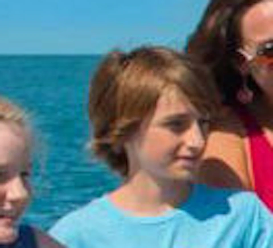 A woman and two children stand on a boat's deck, looking out over the ocean under a clear blue sky.
