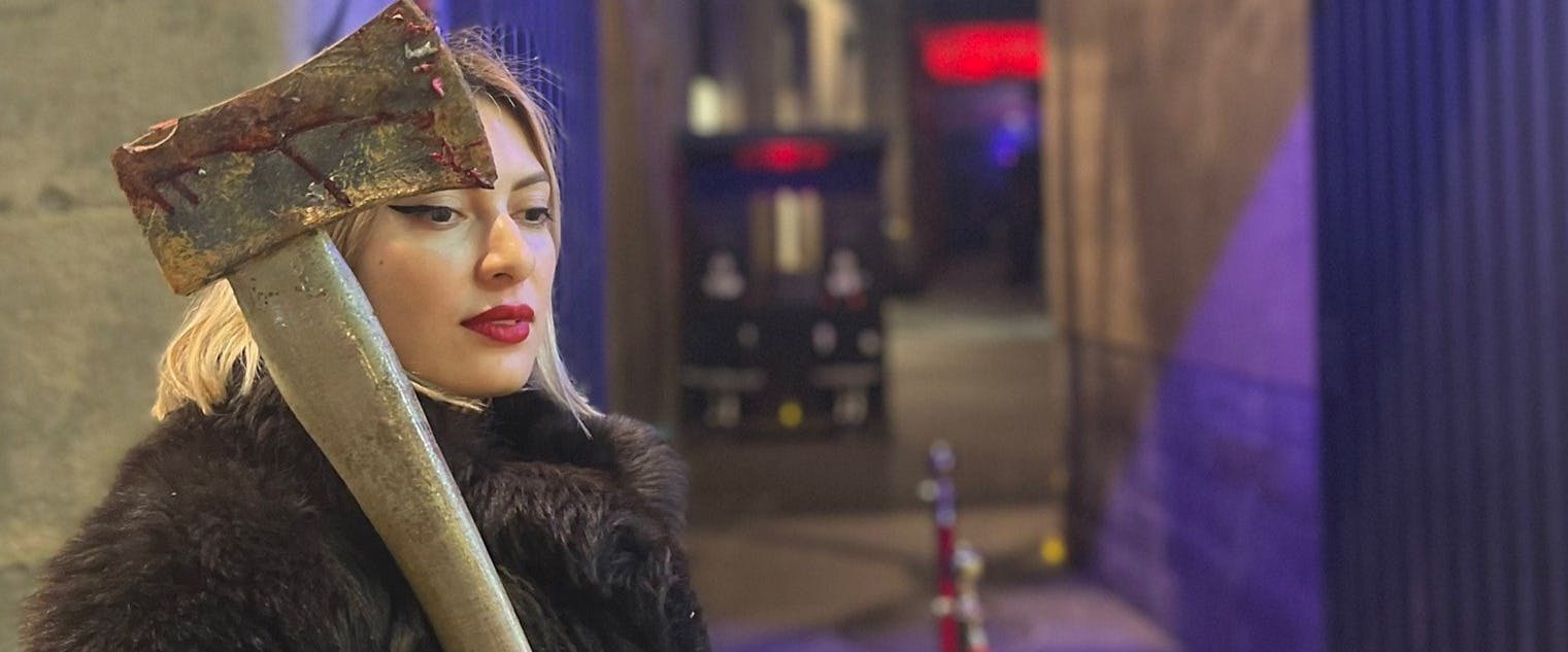 Kobieta z blond włosami i czerwoną szminką trzyma zakrwawiony topór nad ramieniem w słabo oświetlonej miejskiej uliczce.