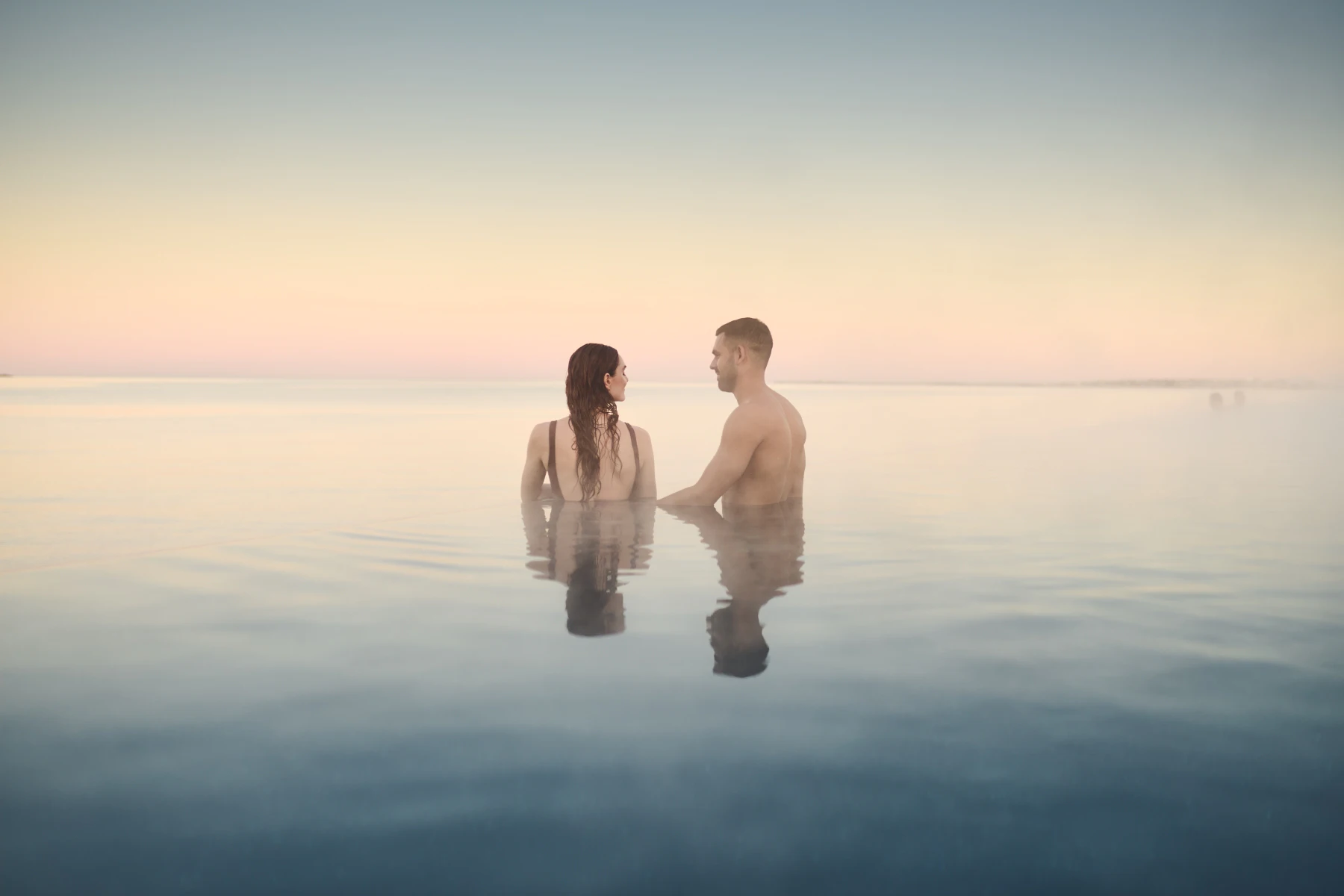 Two people stand in calm water, facing each other during sunrise or sunset, with a clear sky fading from peach to blue.