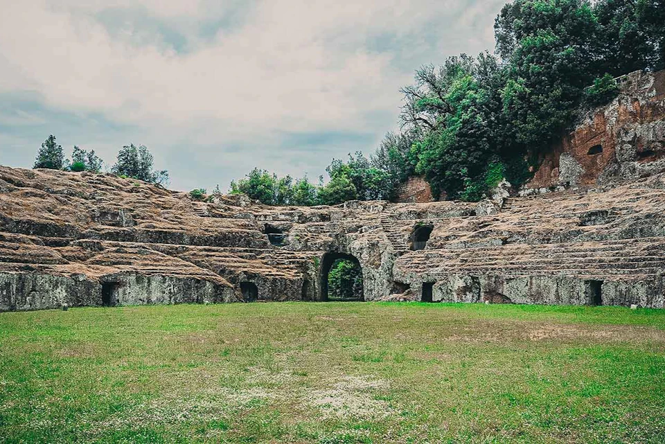 Ancient ruins of an amphitheater with stone arches and greenery, surrounded by grass and trees under a cloudy sky.
