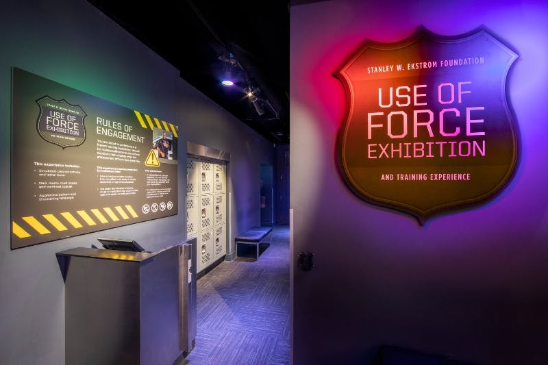 Museum exhibit with a sign reading "USE OF FORCE EXHIBITION." Adjacent wall displays "RULES OF ENGAGEMENT" text. Dim lighting.