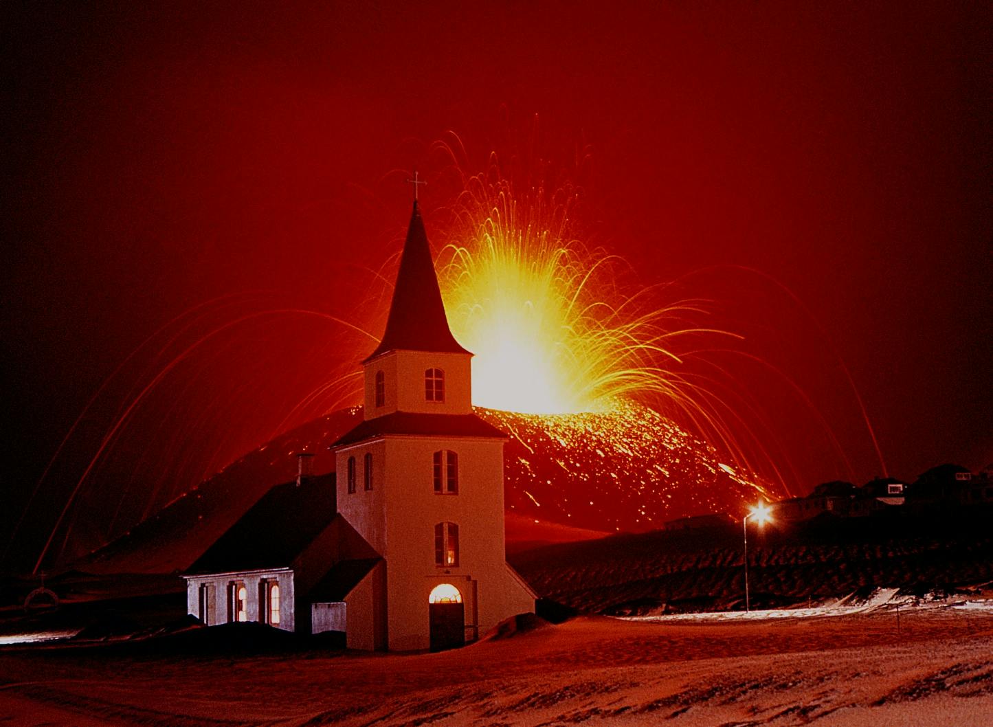 A white church stands in the foreground with an erupting volcano emitting bright lava in the background, under a red sky.