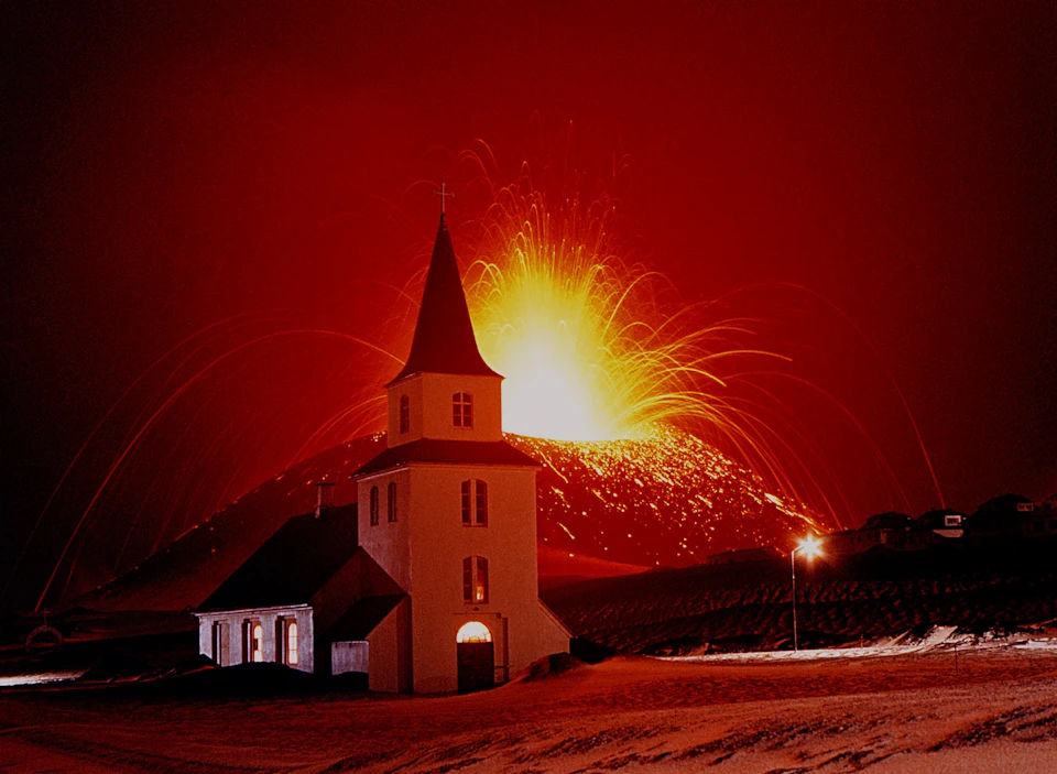 A white church stands in the foreground with an erupting volcano emitting bright lava in the background, under a red sky.