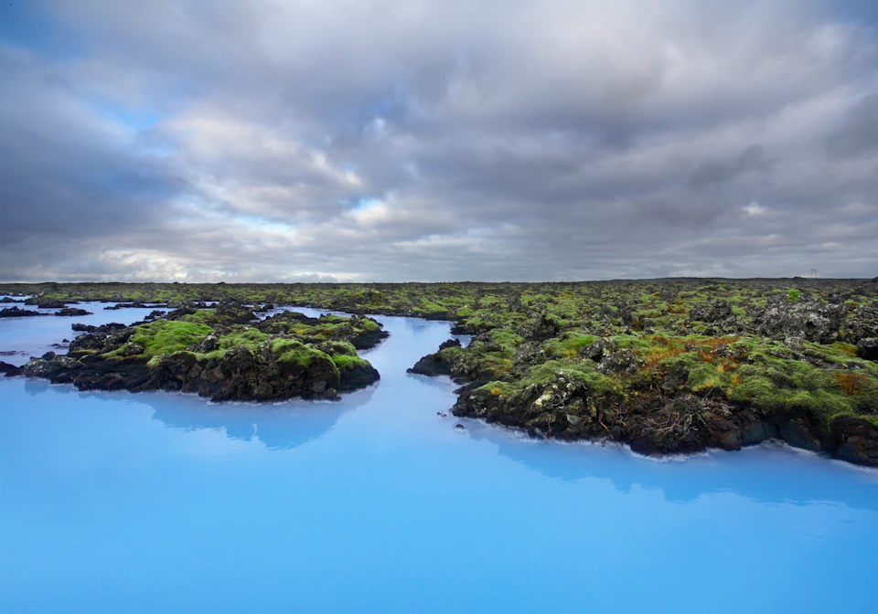 A serene landscape with vivid blue water surrounded by dark moss-covered rocks and a cloudy sky overhead.