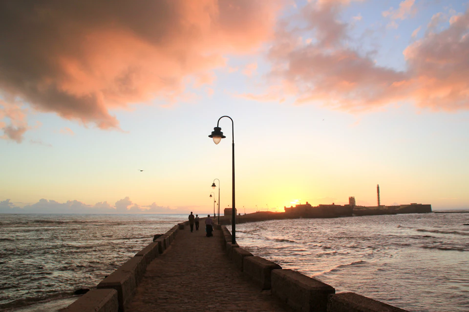 A stone pier with streetlights extends into the sea at sunset, with a lighthouse visible in the distance and a few people walking.