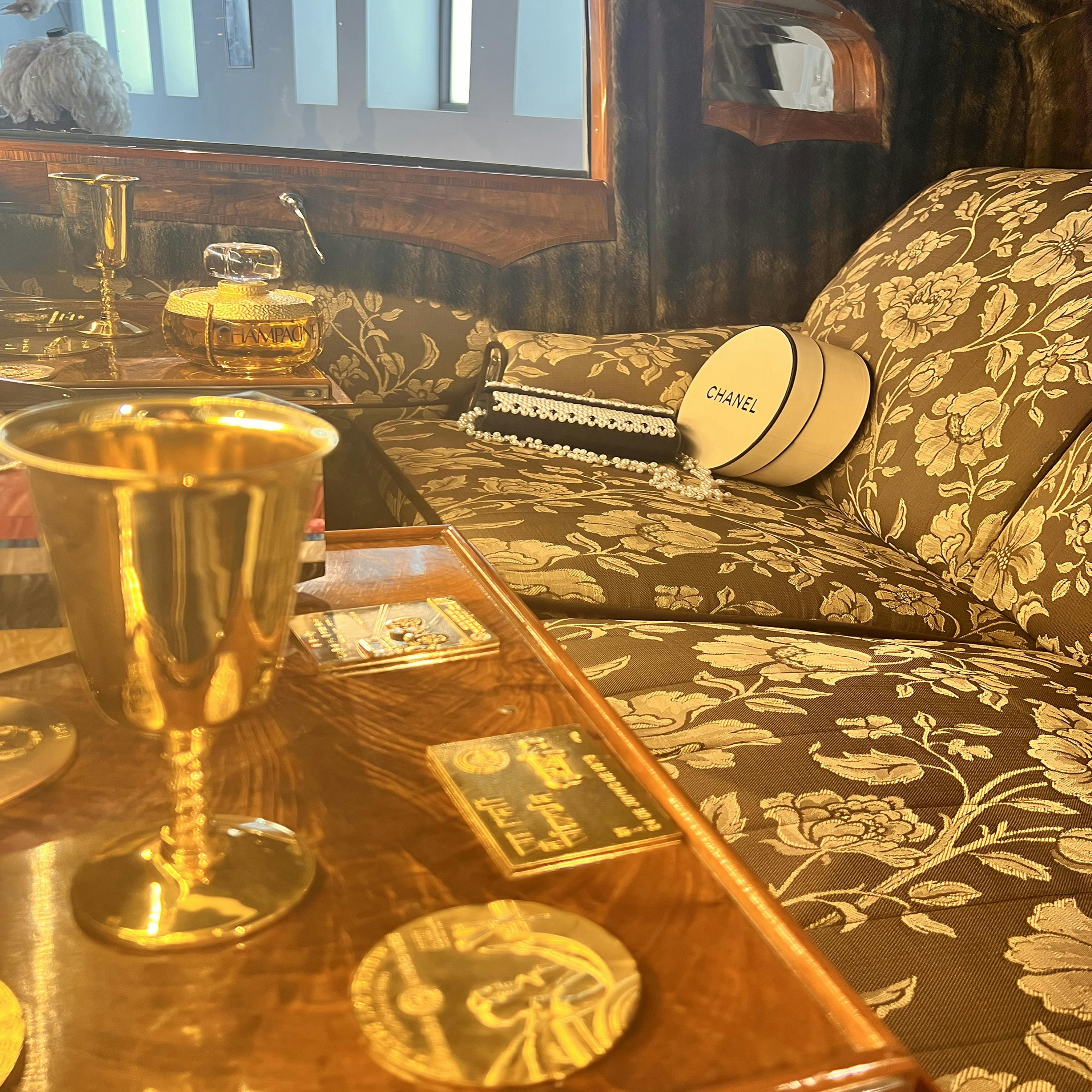 Luksuriøst interiør med blomstermønstrede siddepladser, guldbægre på et træbord og en Chanel-hatteæske på sofaen.