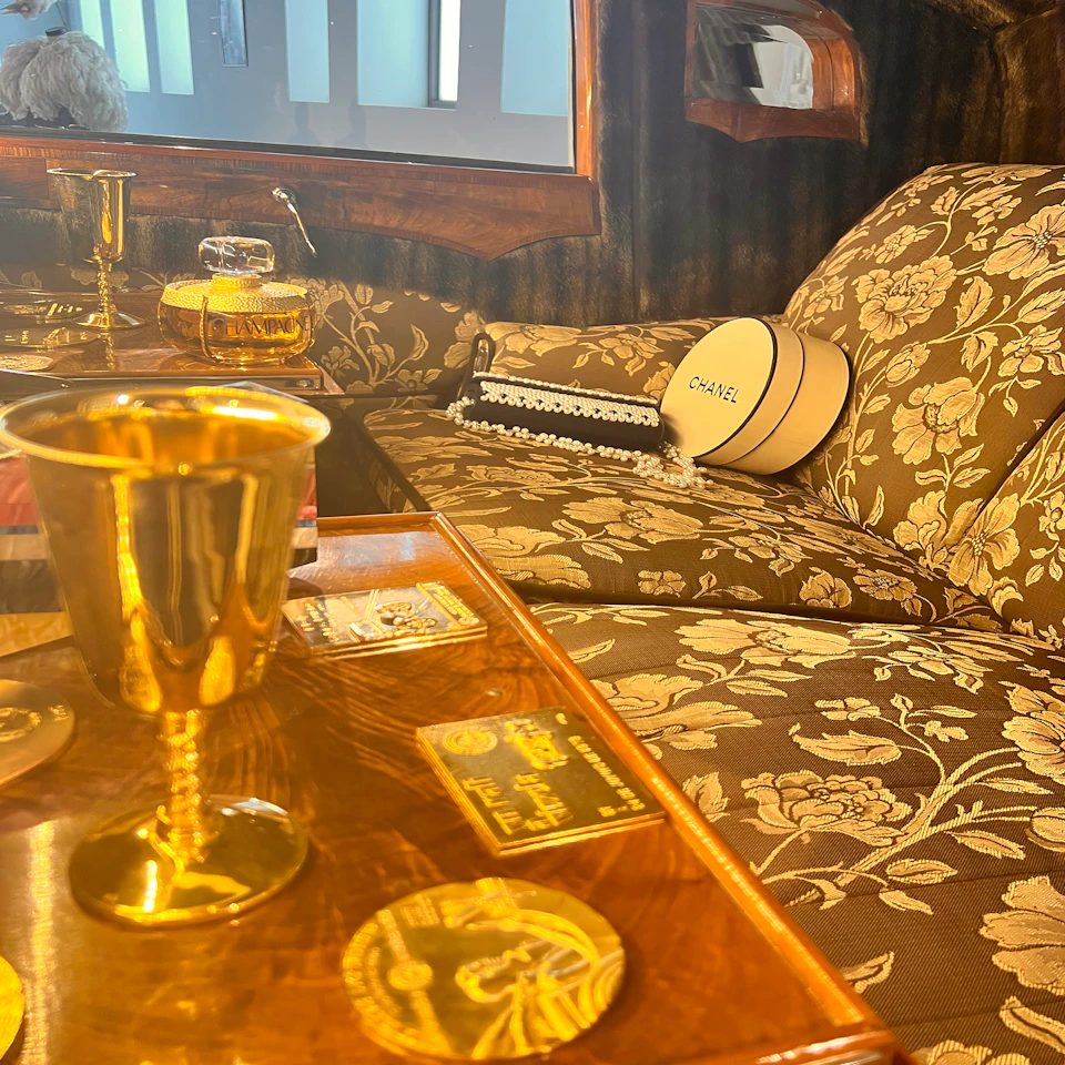 Luxurious interior with floral-patterned seating, gold goblets on a wooden table, and a Chanel hat box on the couch.