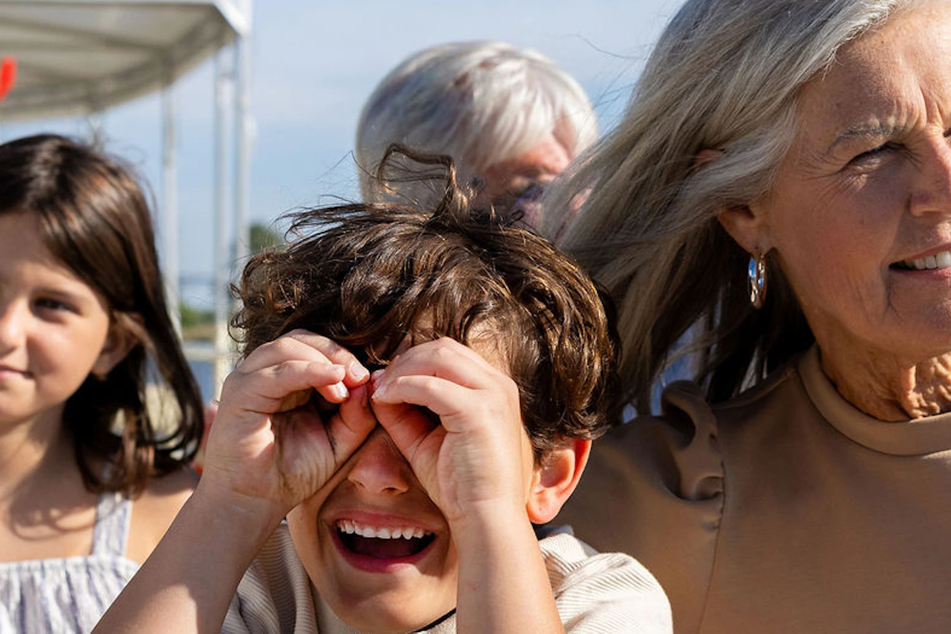 A child using binoculars, a woman smiling, and a girl sitting on a bench outdoors under a sunny blue sky.