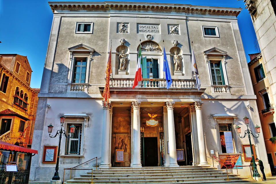 A historic building facade with statues, columns, and multiple flags, including Italian. Latin inscriptions are visible above.
