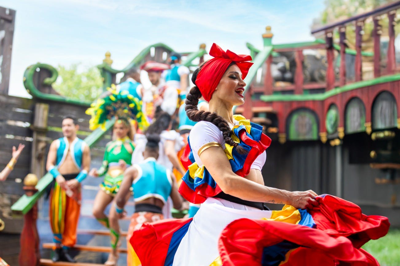 A group of performers in colorful costumes on a stage, with one woman in a red headscarf and traditional dress in focus.