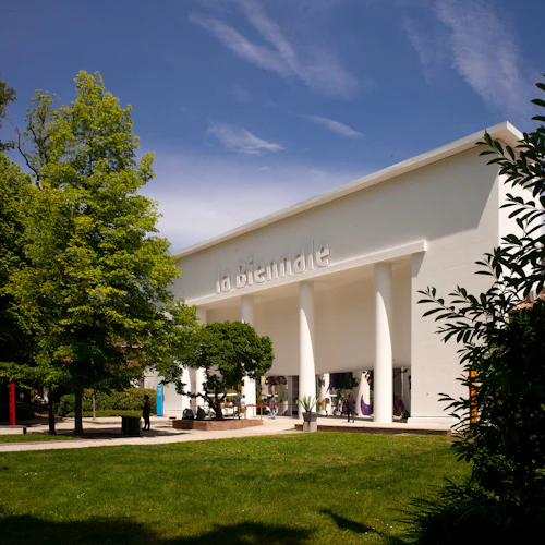 Stor vit byggnad med "la Biennale" skrivet på den, omgiven av träd och grönska under en blå himmel.