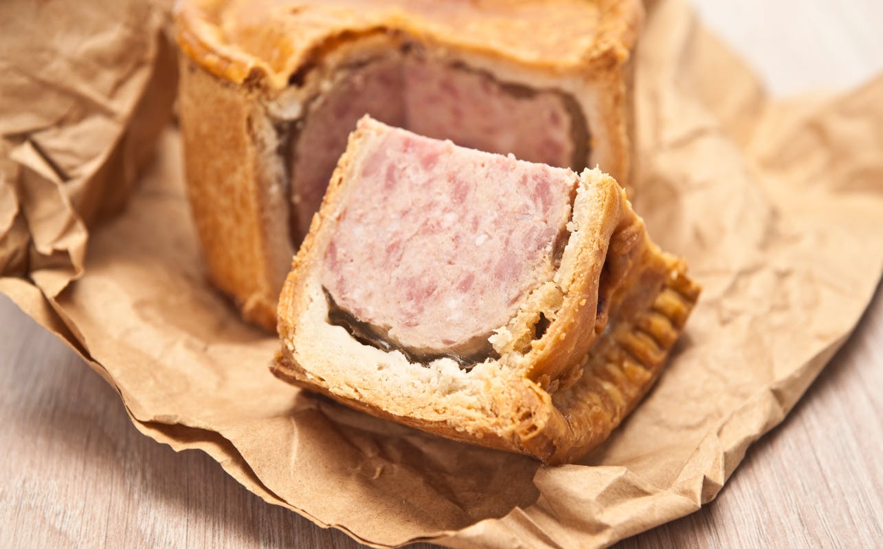 A close-up of a partially cut meat pie on crumpled brown paper. The filling is pink with a brown crust.