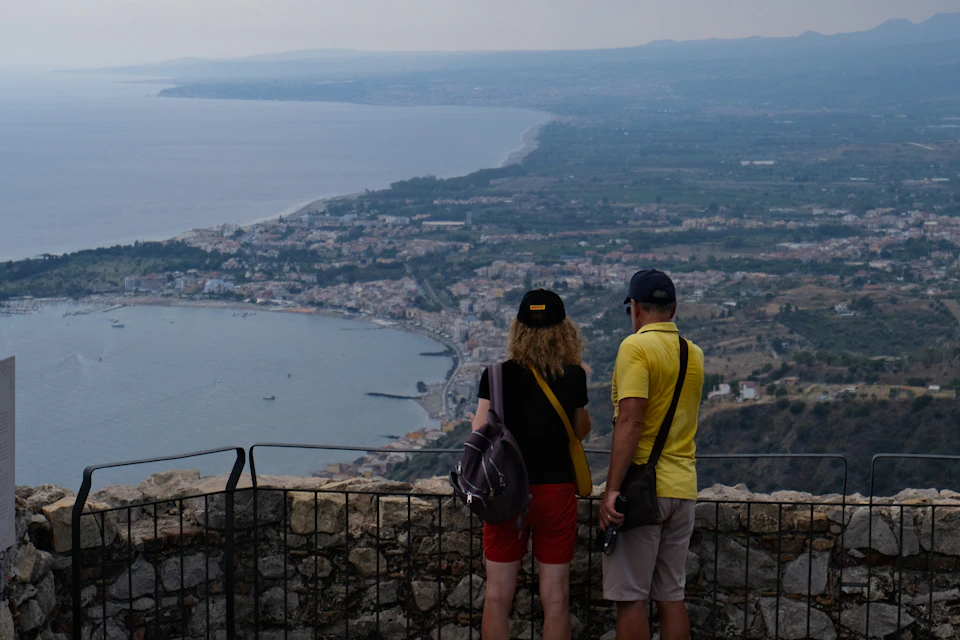 Two people wearing hats and carrying bags stand at a railing, overlooking a coastal town and expansive landscape.