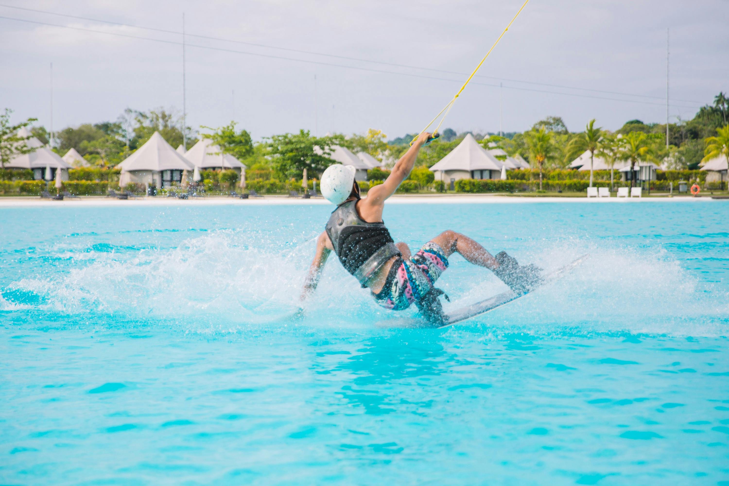 Person wakeboarding on a blue water surface near a resort with tents and greenery in the background.