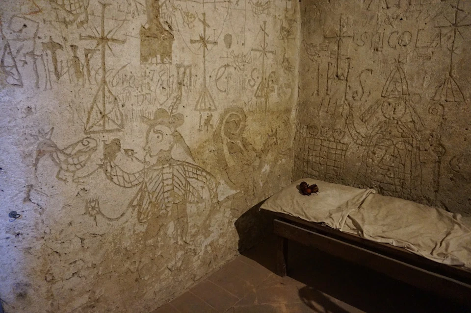 A small room with walls covered in old drawings and carvings; a simple bed with worn linens and a bundle of red fabric on it.