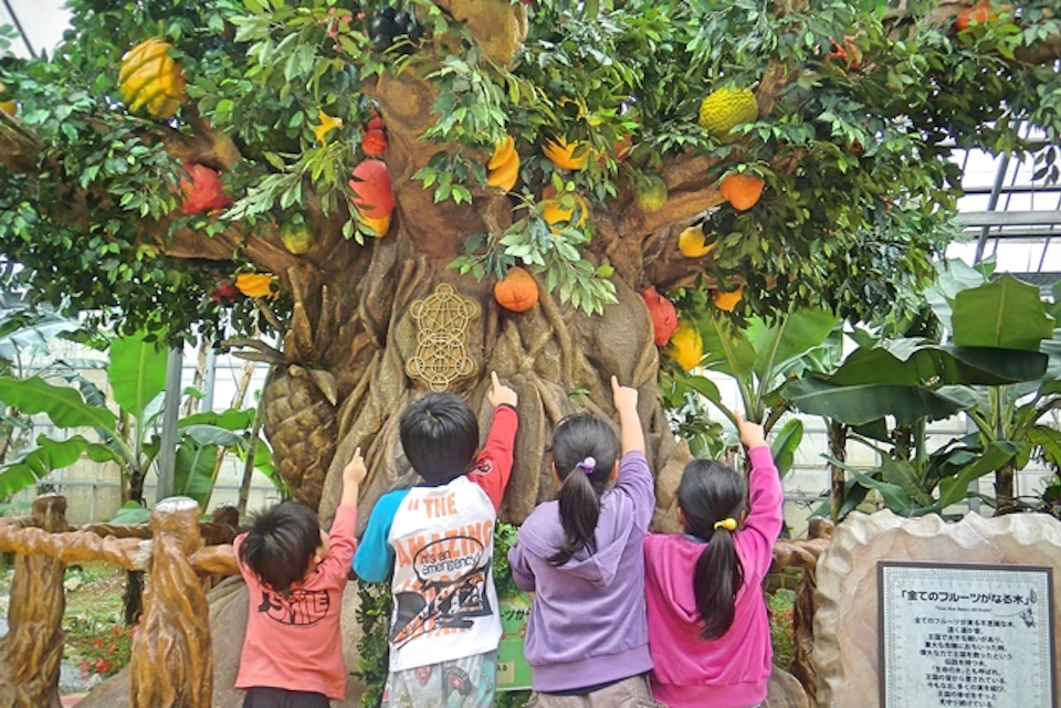 Four children pointing at a colorful artificial tree with fruit decorations in a green indoor environment.