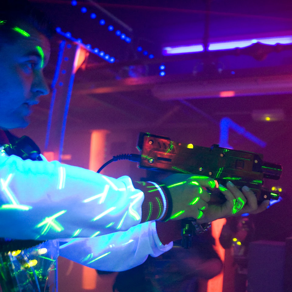 Person wearing a white jacket with luminous designs, holding a laser tag gun in a dark, neon-lit environment.