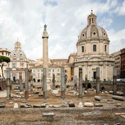Forntida ruiner med pelare, en hög staty-toppad pelare och två kupolformade kyrkor i bakgrunden under en molnig himmel.