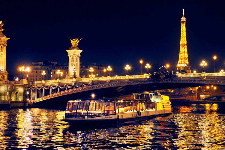 A nighttime view of a lit-up bridge, tour boat on the river, and the Eiffel Tower in the background in Paris.