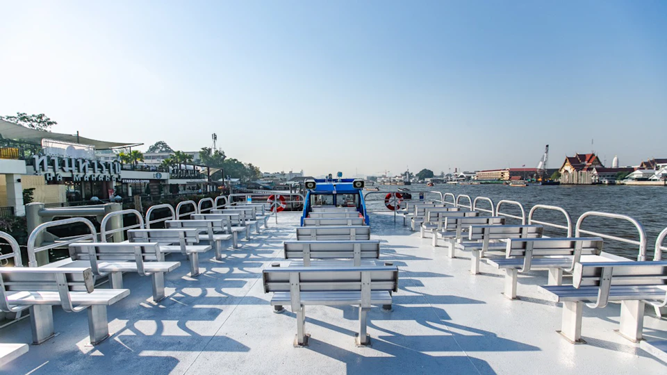 Empty upper deck of a ferry with rows of benches, life buoys on the sides, and a calm river with buildings and trees in the background.
