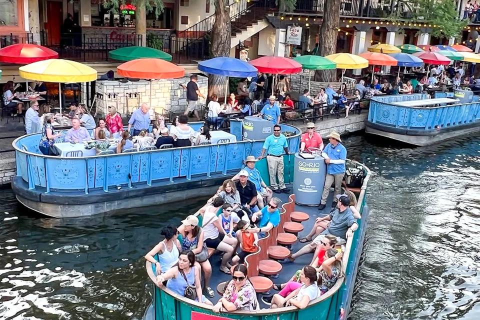 People enjoying a boat ride on a waterway surrounded by restaurants with colorful umbrellas and vibrant activity.