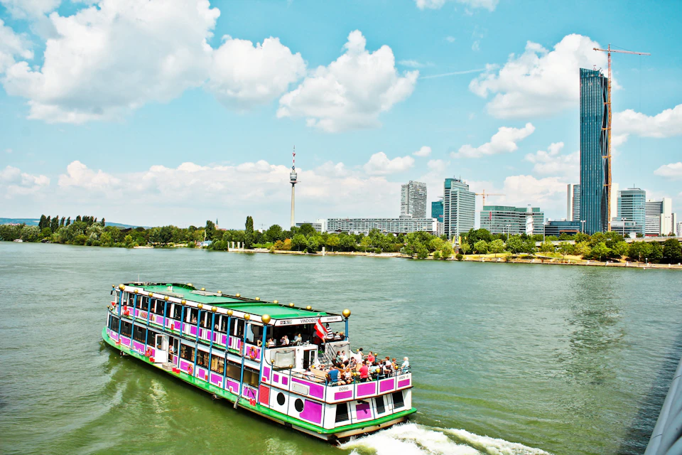 A multi-deck riverboat with passengers cruises a river, with a city skyline and a tall tower visible in the background under a blue sky.