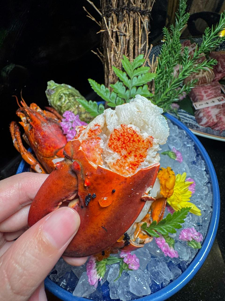 A hand holding a cooked lobster claw with meat exposed, surrounded by decorative greenery and flowers on a platter.