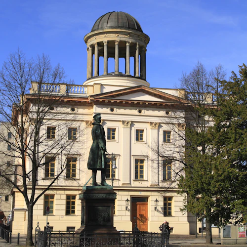 Bronsstaty av en man framför en neoklassisk byggnad med ett välvt torn, omgivet av träd och en kullerstensplaza.