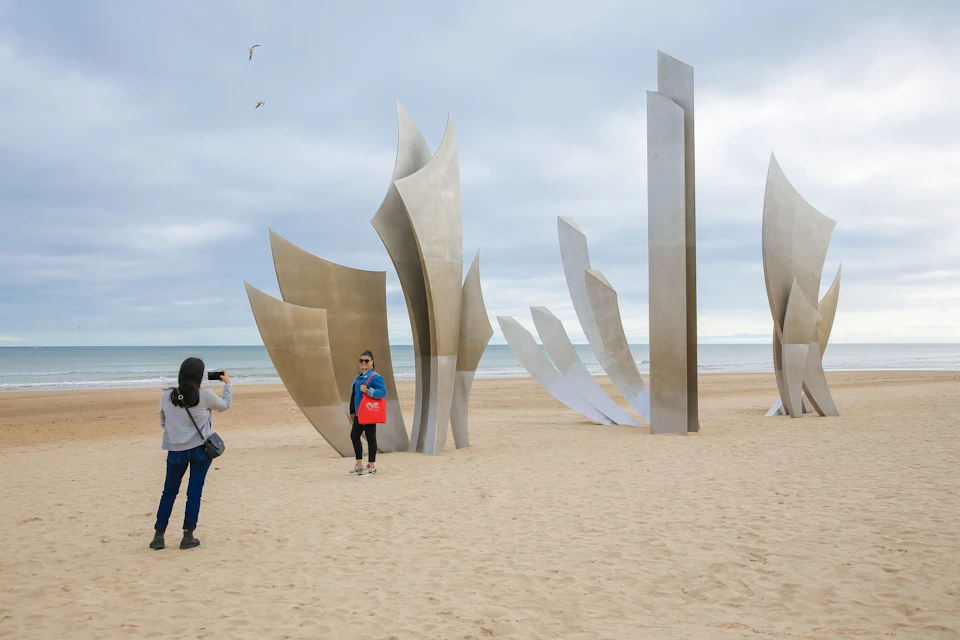 People taking photos near large metal sculptures on a sandy beach with the ocean and cloudy sky in the background.