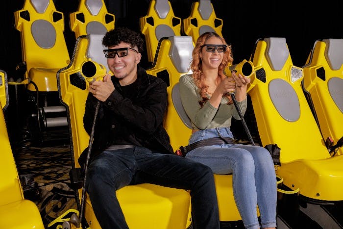 Two people wearing 3D glasses and holding laser guns sit in yellow seats in a dark room, smiling and enjoying the ride.