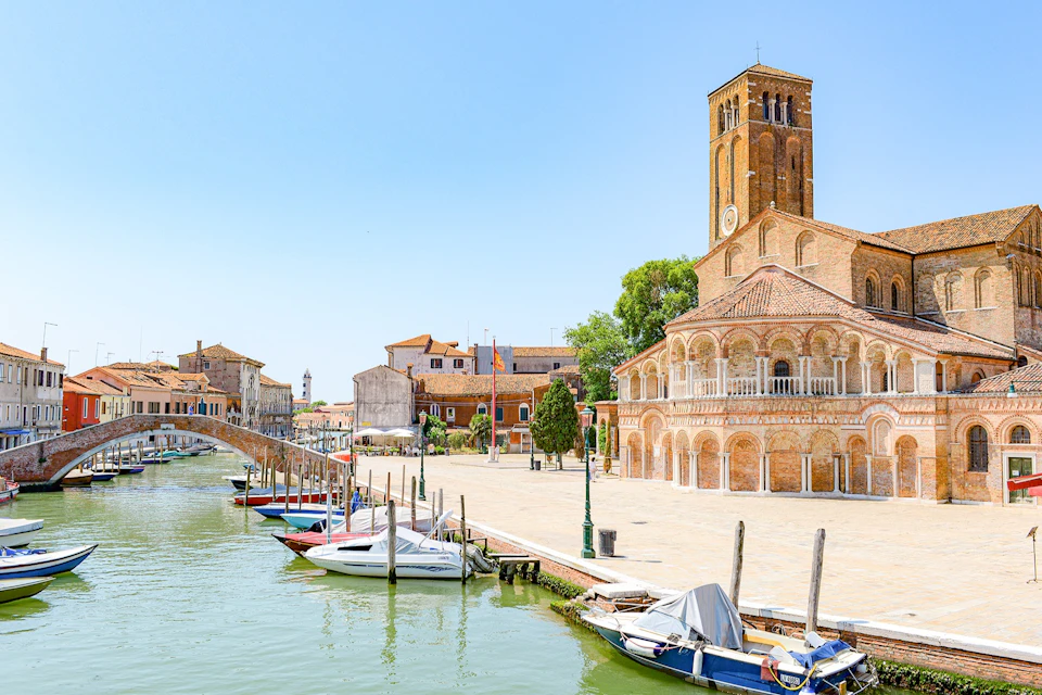 A canal with boats, a stone bridge, and an ornate brick church in a sunny square in a European-style town.