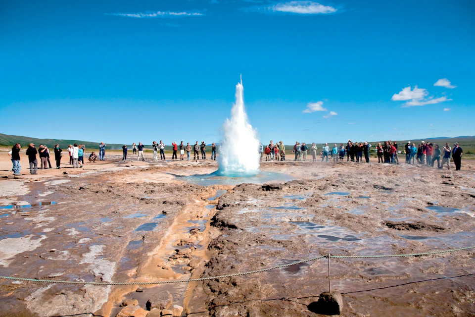 People observing a geyser erupting under a clear blue sky, surrounded by rocky terrain and sparse greenery in the background.