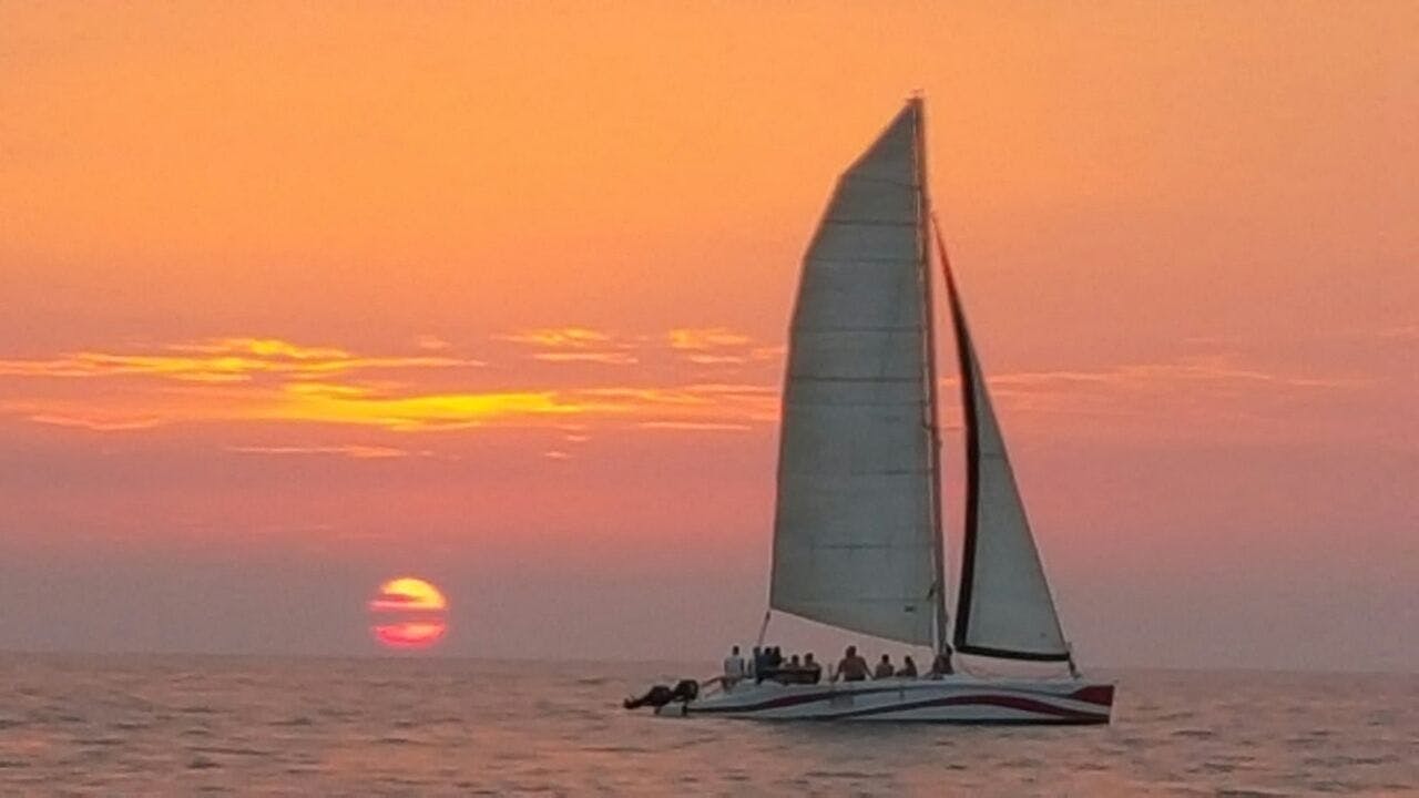 Catamaran sailboat with several people aboard on a calm sea at sunset, with the sun partially below the horizon.