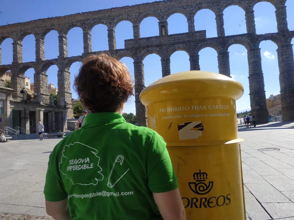 Person in a green shirt stands near a yellow Correo mailbox, with an ancient aqueduct in the background on a sunny day.