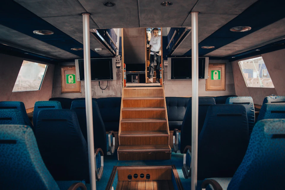 Interior of a vehicle or vessel with blue seats, a wooden staircase, and a person standing at the top of the stairs.