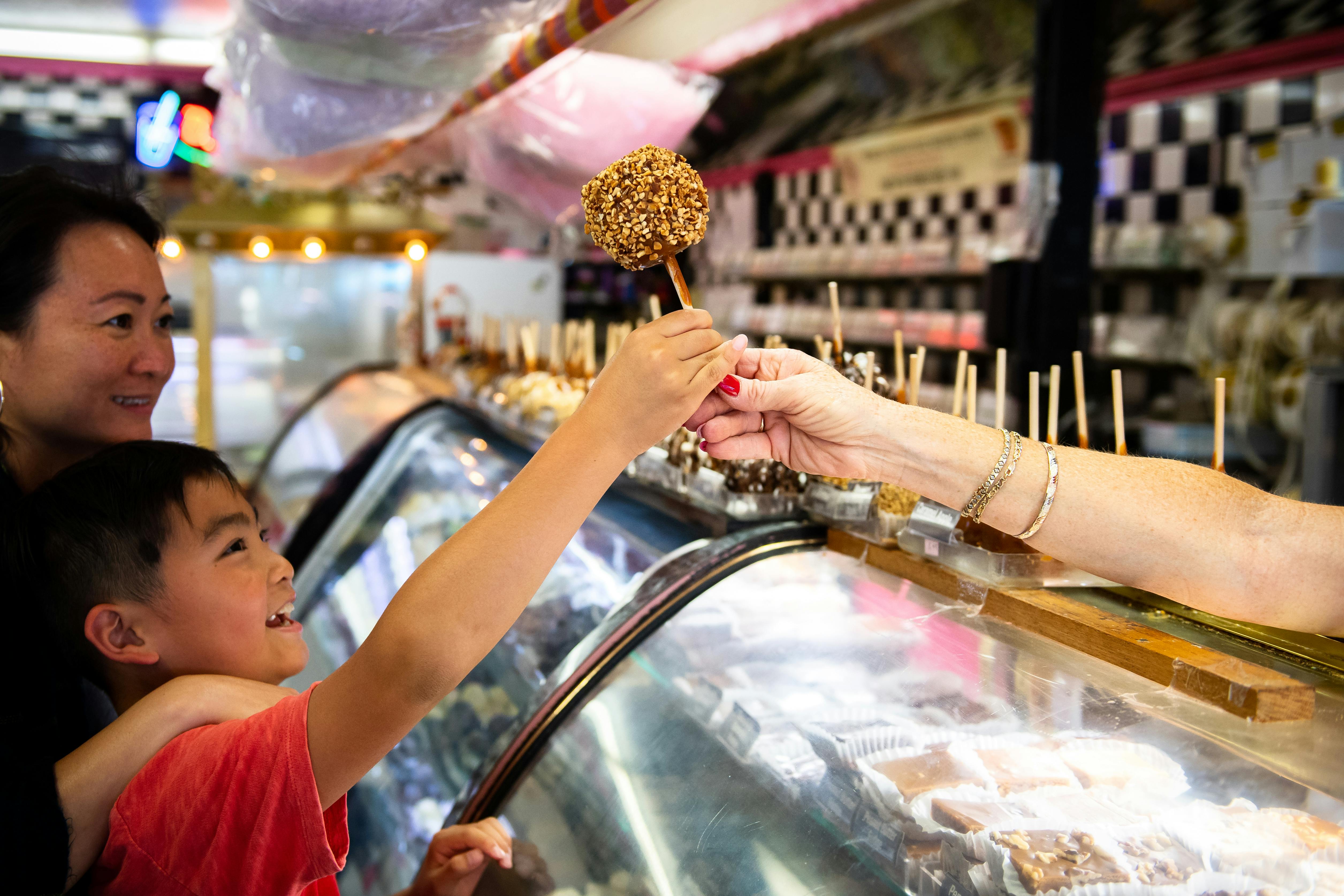 Ένα χαμογελαστό αγόρι λαμβάνει ένα μήλο καραμέλας σε ένα ραβδί από έναν πωλητή, ενώ μια γυναίκα στέκεται δίπλα του σε ένα ζαχαροπλαστείο.