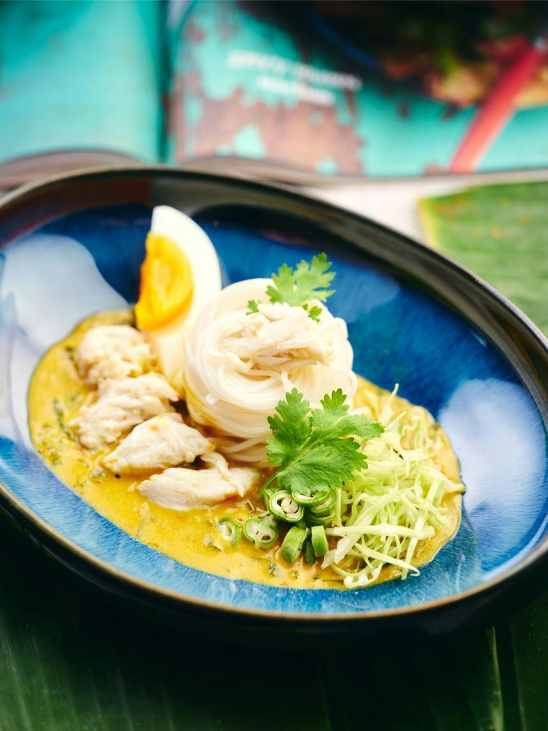 A bowl of noodles in yellow curry sauce, topped with chicken, egg, cilantro, and vegetables, served in a blue dish.