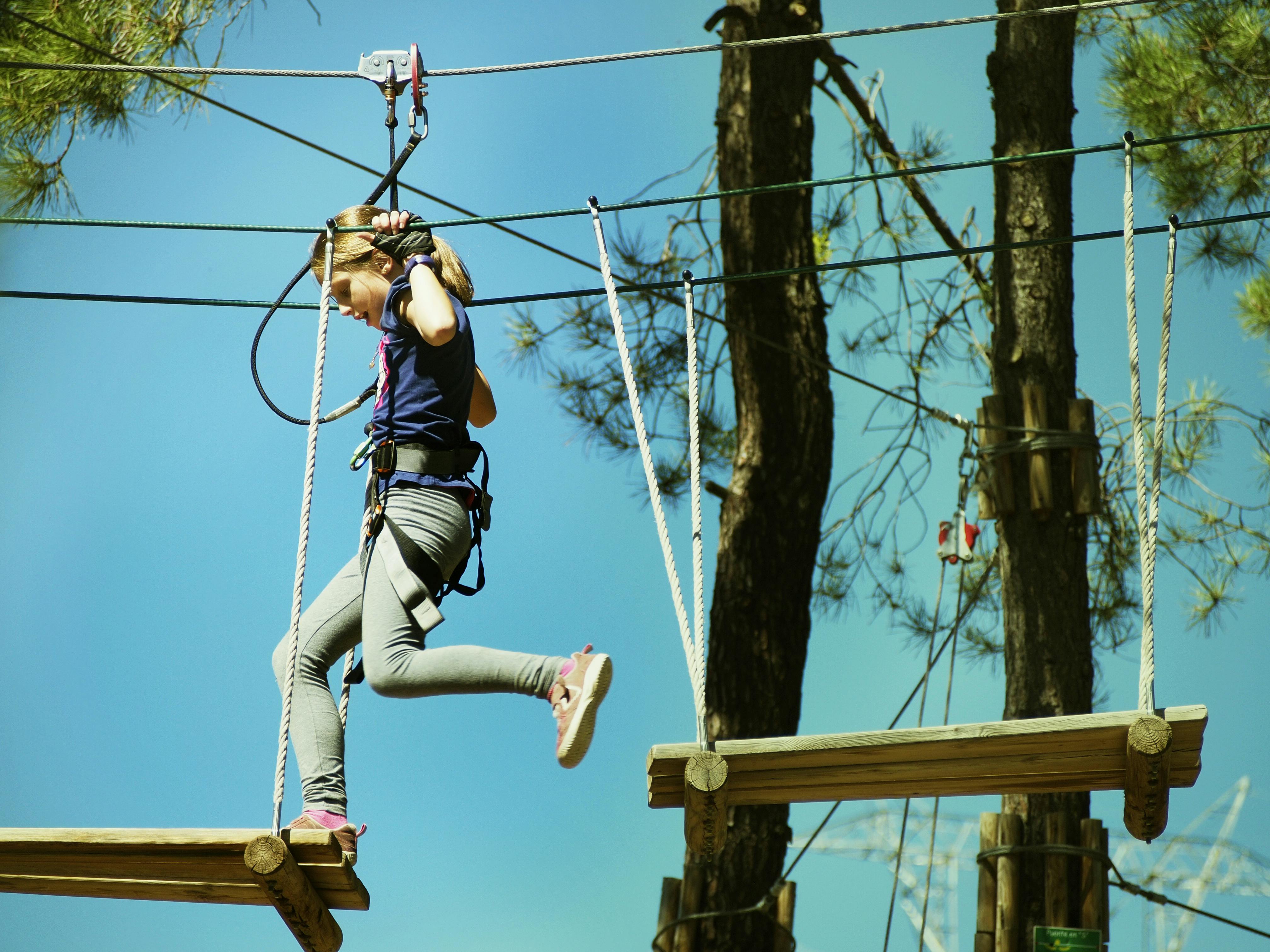 A girl wearing a harness crossing a wooden platform on a high ropes course, surrounded by trees and a clear blue sky.