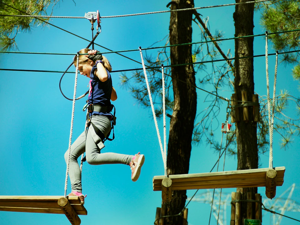 A girl wearing a harness crossing a wooden platform on a high ropes course, surrounded by trees and a clear blue sky.