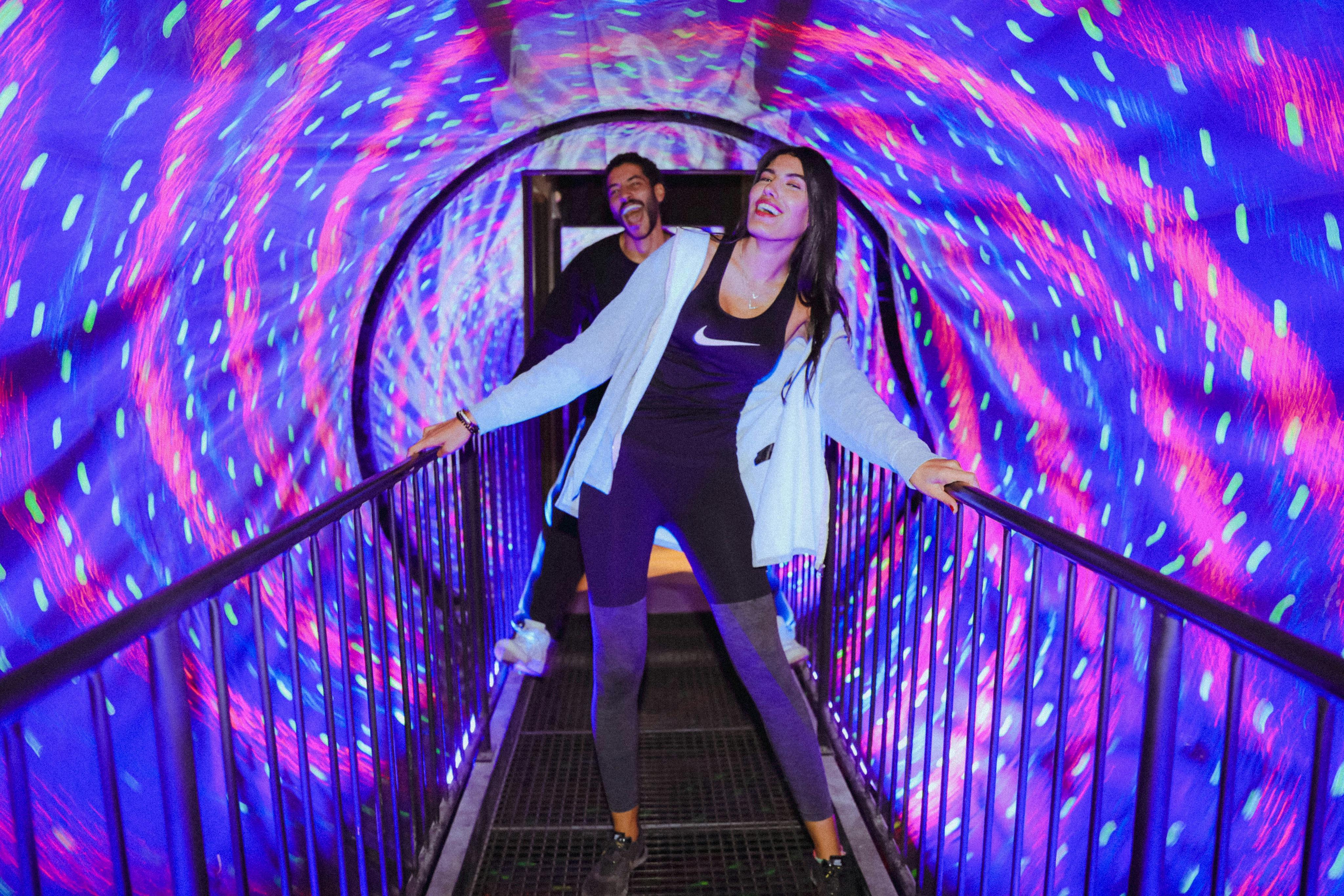 Due persone si godono un tunnel vorticoso e colorato, con luci vivaci che creano un effetto vorticoso intorno a una stretta passerella.