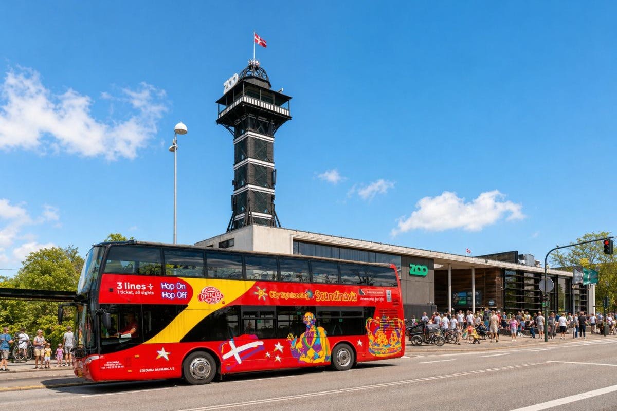 En rød dobbeltdækker tourbus foran en zoologisk indgang med et højt observationstårn i baggrunden.