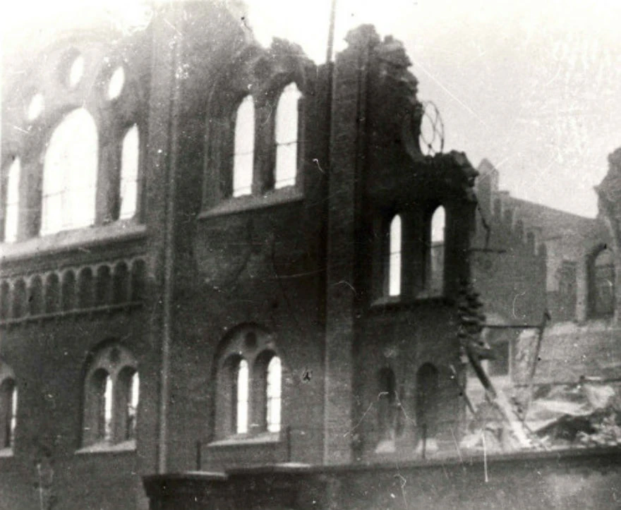 Damaged brick building with arched windows, partial collapse, and rubble. Scene appears old and monochrome.