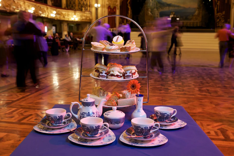 An elegant tea setup with a tiered tray of snacks and tea cups in a grand hall with blurred people in the background.